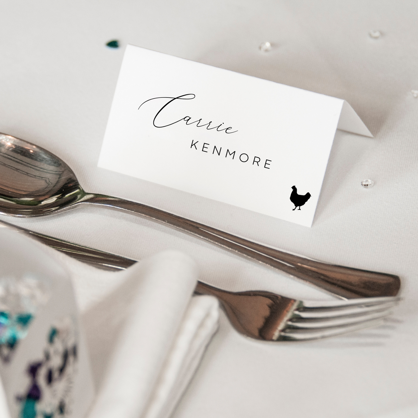 A white place card can be seen on a white tablecloth with cutlery in front. The place card reads "Carrie Kenmore" with the silhouette of a chicken in the bottom right hand corner.