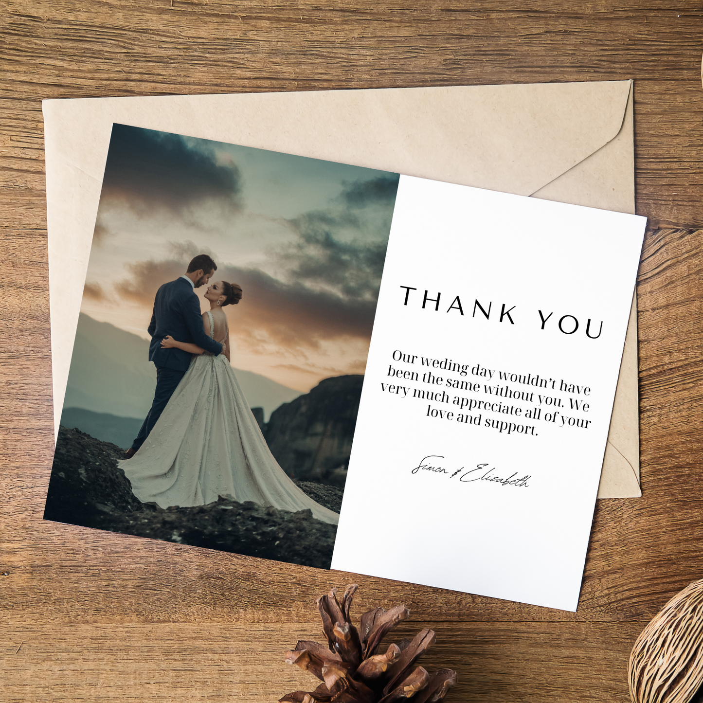 A wedding thank you card lays on top of a light brown envelope on a wooden background. A pinecone can be seen peaking out from the bottom of the image.