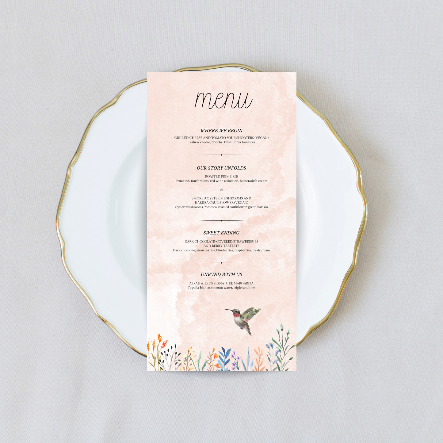 A light pink wedding menu with flowers and a hummingbird at the bottom sits on a white plate with gold trim against a cream coloured background.