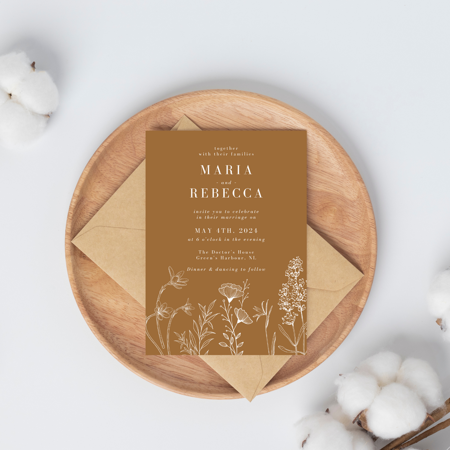 A light brown coloured wedding invitation is placed on top of a beige-coloured envelope on a wodden plate. The plate lays on a white background with clusters of cotton in opposite corners.