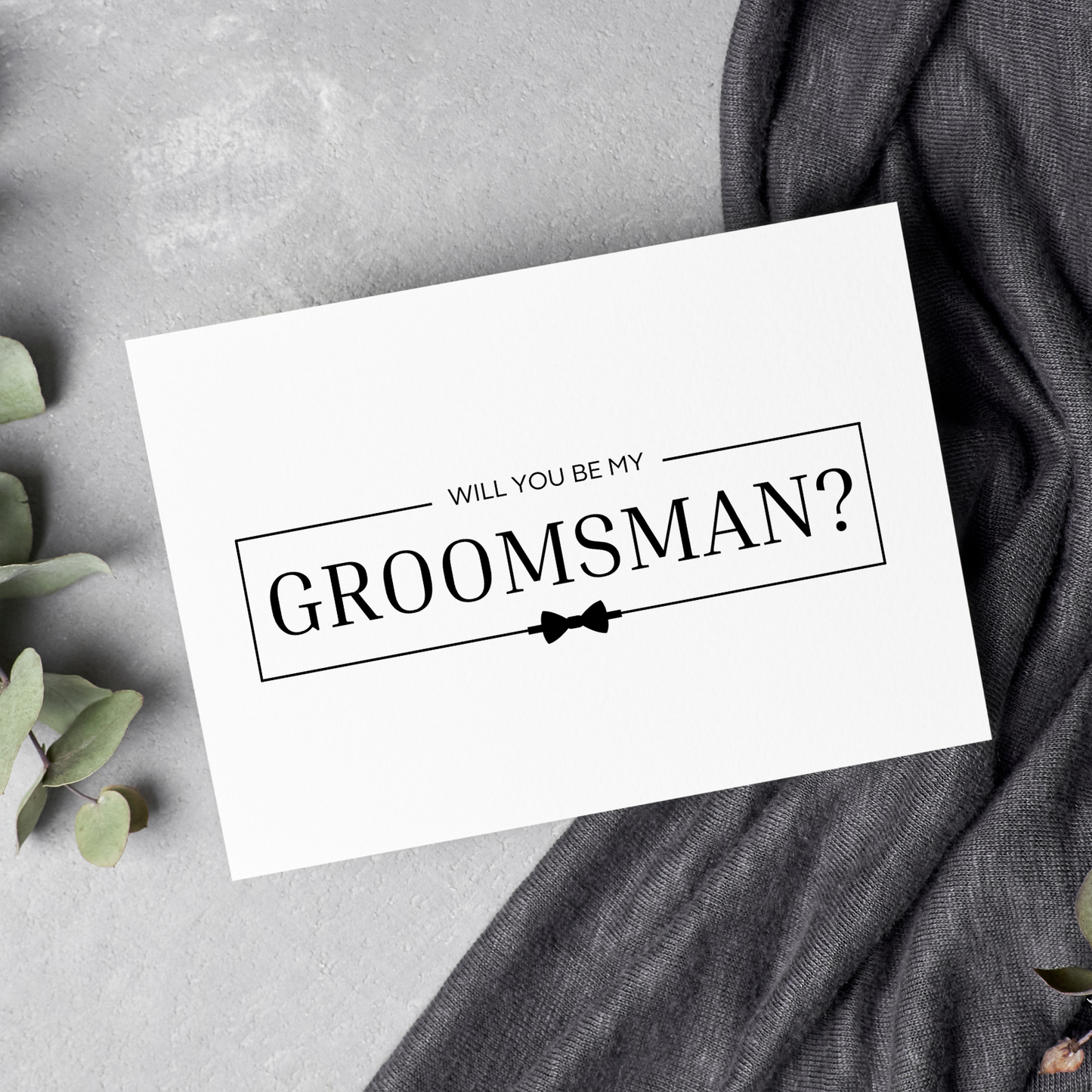 A white card with the phrase "Will you be my groomsman?" can be seen laying against a grey, concrete background. There is a black bow tie under the wording and a darker grey cloth under one half of the card that spans the full height of the image. On the left hand side, a branch of eucalyptus lays adjacent to the card.