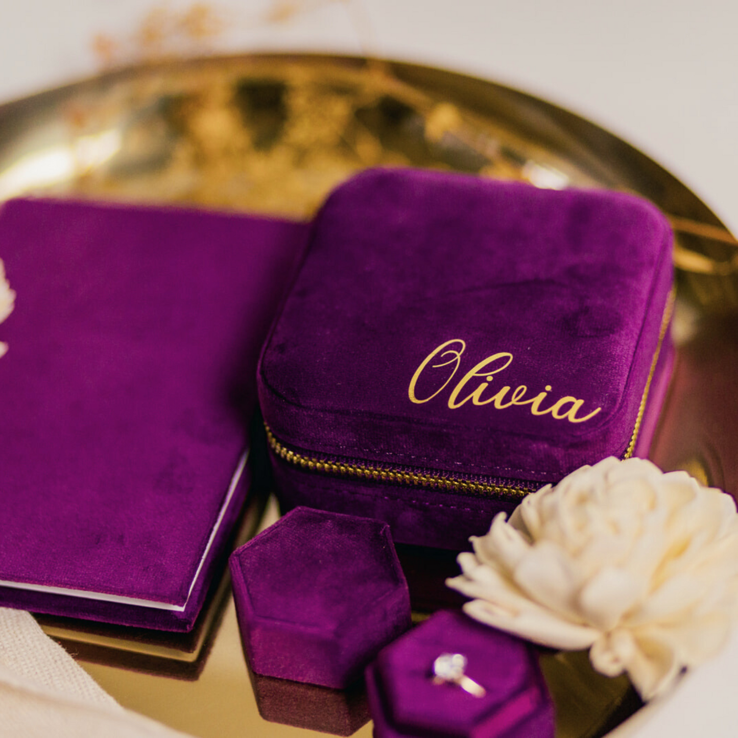 A dark purple jewelry case, vow book and ring box sit in a shallow gold bowl. An off white flower is nestled below the jewelry case which reads "Olivia" in a cursive font.