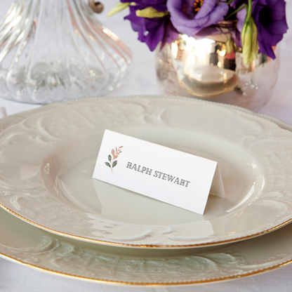 A white place card sits on an ivory coloured plate with the name "Ralph Stewart". There are flowers in a gold vase in front of the place setting and a small illustration of a flower on the placecard.
