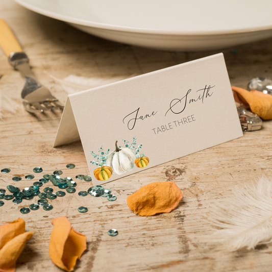 A white place card with a variety of pumpkins in the bottom left hand corner can be seen at a place setting with orange flower petals around it. The place card says "Jane Smith; Table Three".