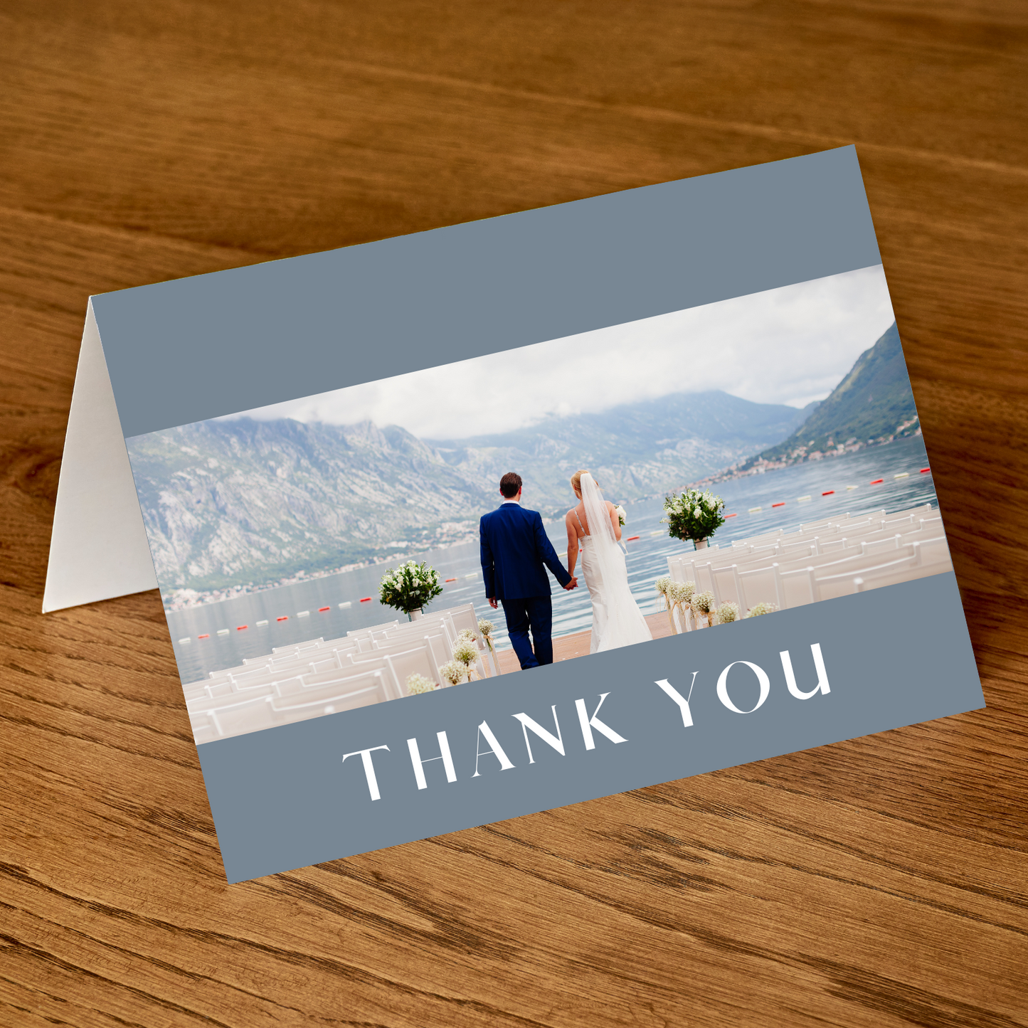 A tented, dusty blue thank you card with a photo of a couple on the front sits on a wooden table.