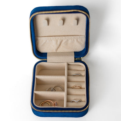 The inside of a sapphire jewelry case. There are three storage sections and seven ring slots on the bottom portion of the case. On the top half, there are three hooks for chains or necklaces and well as a pouch to tuck them in.
