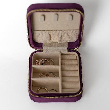 The inside of a plum-cloured jewelry case. There are three storage sections and seven ring slots on the bottom portion of the case. On the top half, there are three hooks for chains or necklaces and well as a pouch to tuck them in.