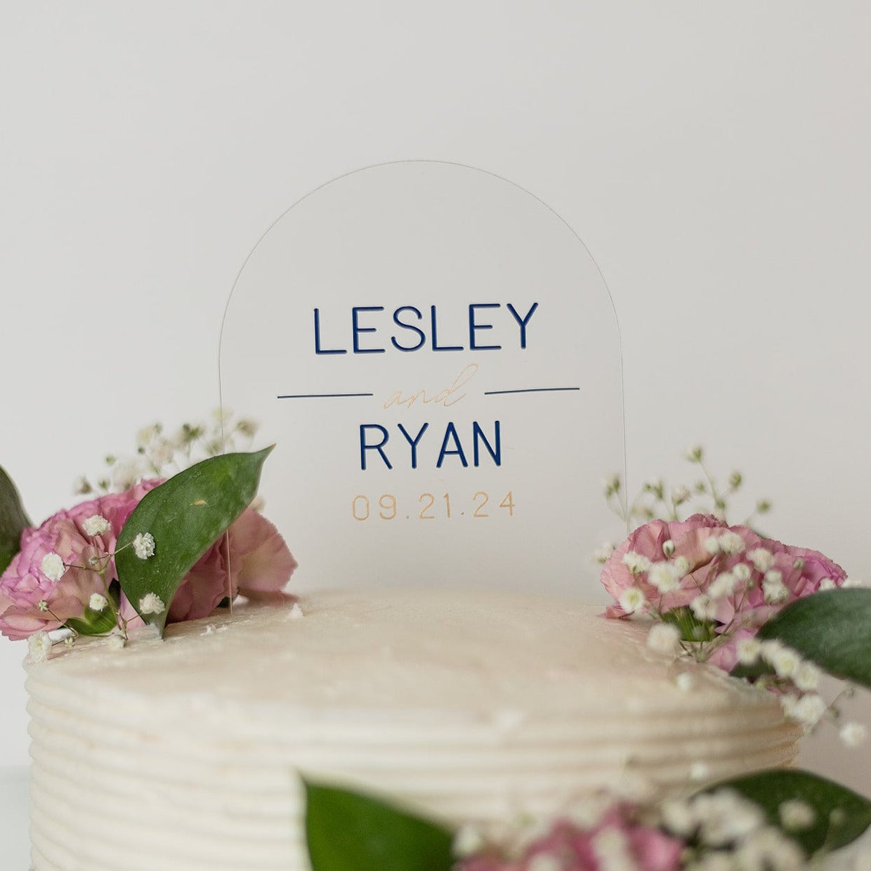 A clear, arch shaped cake topper sits on top of a white cake with pink flowers, sprigs of baby's breath and dark green leaves. The cake topper reads "Lesley and Ryan, 09.21.24".