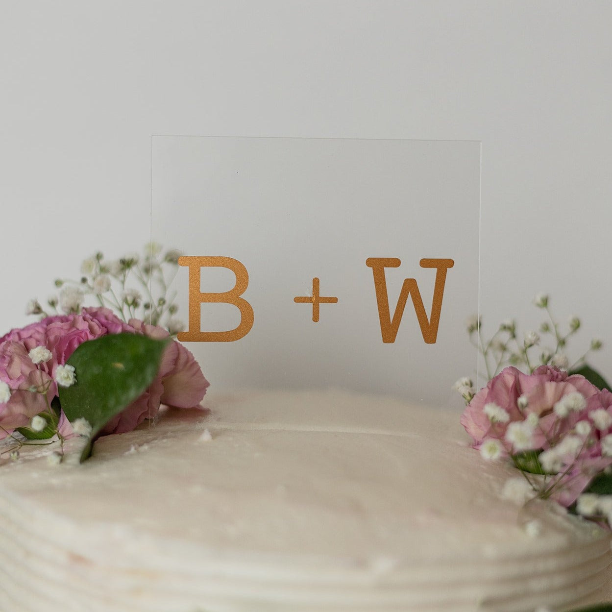 A clear, square shaped cake topper sits on top of a white cake with pink flowers, sprigs of baby's breath and dark green leaves. The cake topper reads "B + W"