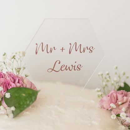 A clear, hexagon shaped cake topper sits on top of a white cake with pink flowers, sprigs of baby's breath and dark green leaves. The cake topper reads "Mr. + Mrs. Lewis".