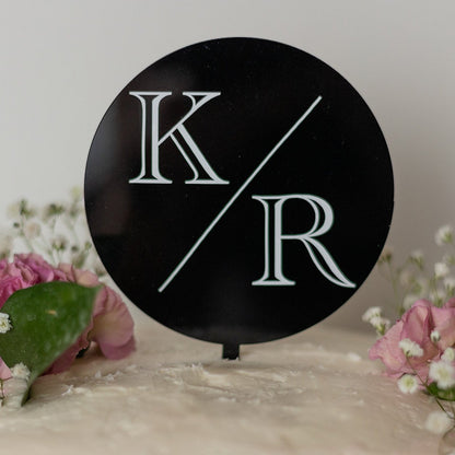 A black circle shaped cake topper sits on top of a white cake with pink flowers, sprigs of baby's breath and dark green leaves. The cake topper has the initials "K" and "R" separated by a long diagonal line.