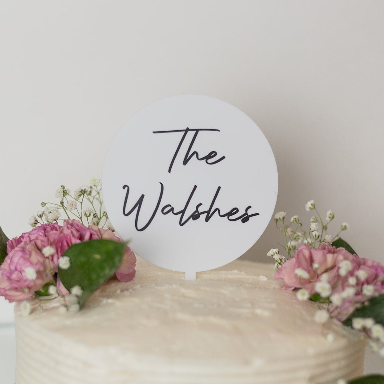 A white circle shaped cake topper sits on top of a white cake with pink flowers, sprigs of baby's breath and dark green leaves. The cake topper reads "The Walshes"