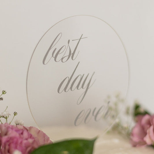 A clear, circle shaped cake topper sits on top of a white cake with pink flowers, sprigs of baby's breath and dark green leaves. The cake topper reads "best day ever".