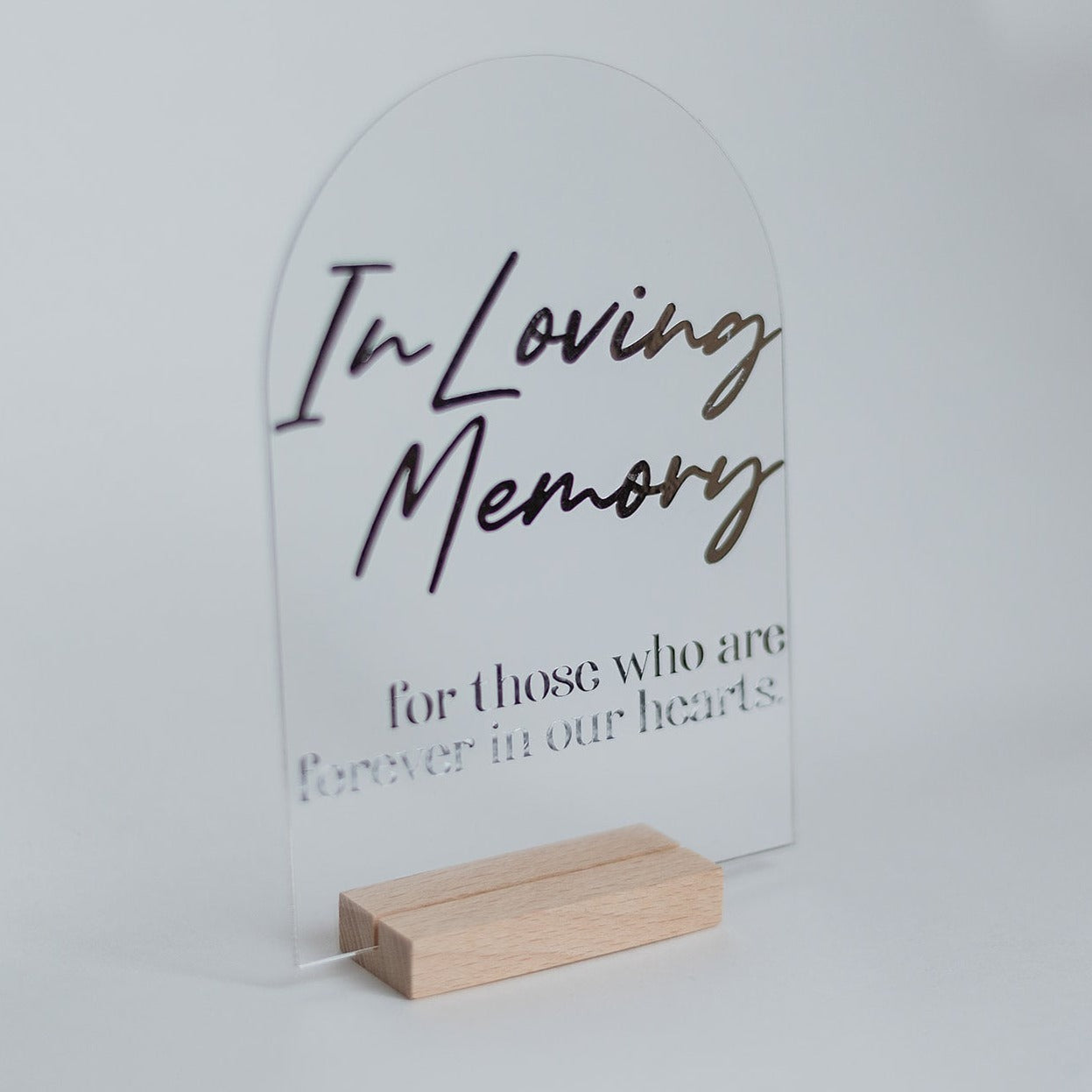 A clear, arch-shaped acrylic memorial sign with silver text on a light wooden base.