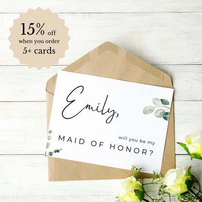 A white card with the phrase "Emily, will you be my maid of honour" is seen on top of a kraft paper envelope. The card and envelope are laying on a light-coloured wood backdrop with yellow roses in the bottom right hand corner. A beige 'burst' shape can be seen in the top left hand corner with "15% off when you order 5+ cards" inside.