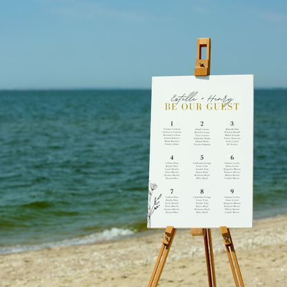 A seating chart sits on a wooden easel at a beach. The chart reads "Estelle + Henry... be our guest". A list of tables and guests can be seen in the middle with the outline of a flower at the bottom left hand corner of the chart.