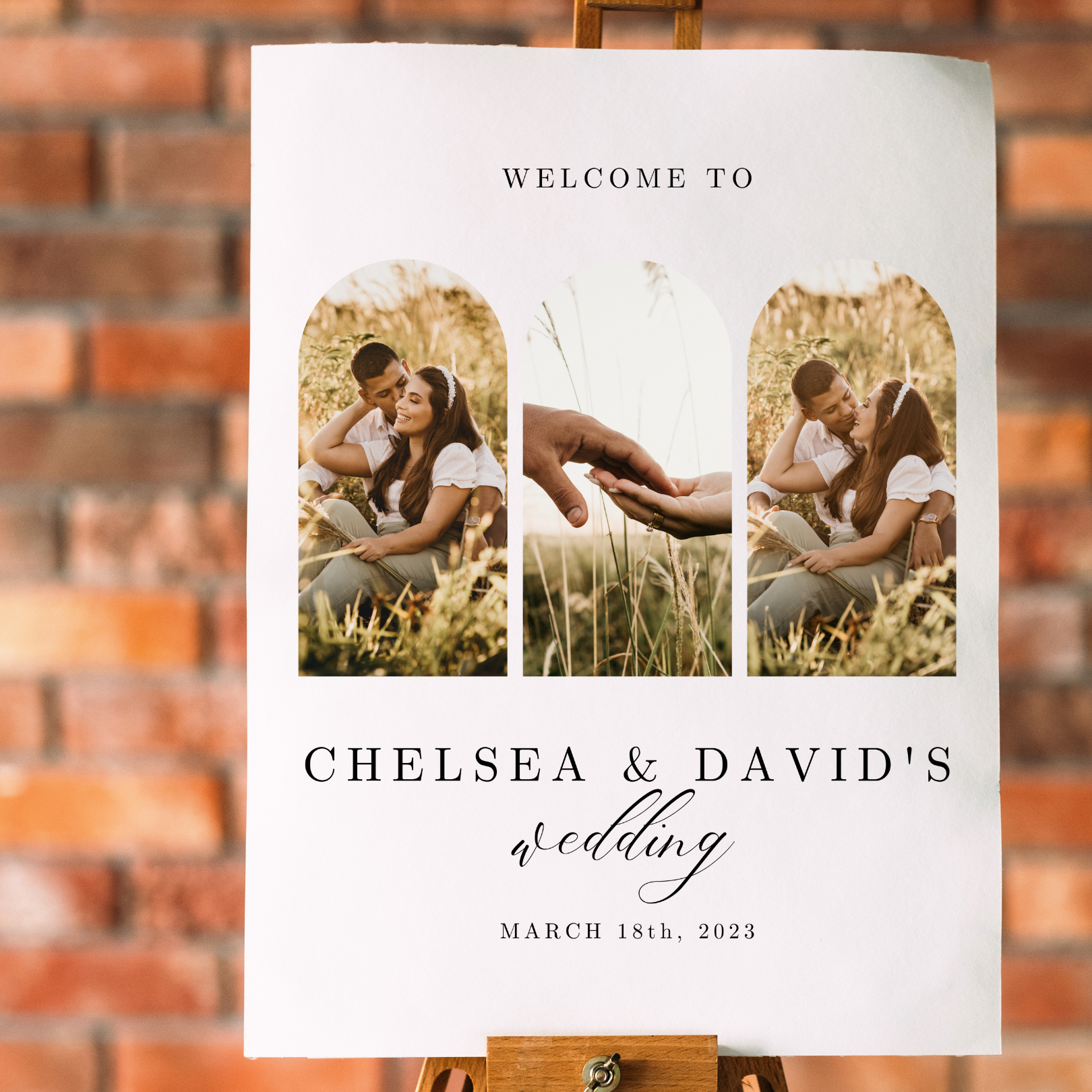 A wedding welcome sign with pictures of a couple in a grassy field sits on an easel against a brick background. 