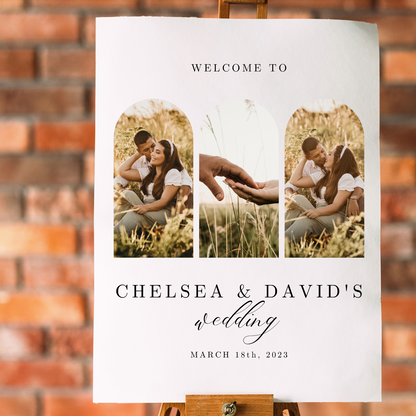 A wedding welcome sign with pictures of a couple in a grassy field sits on an easel against a brick background. 