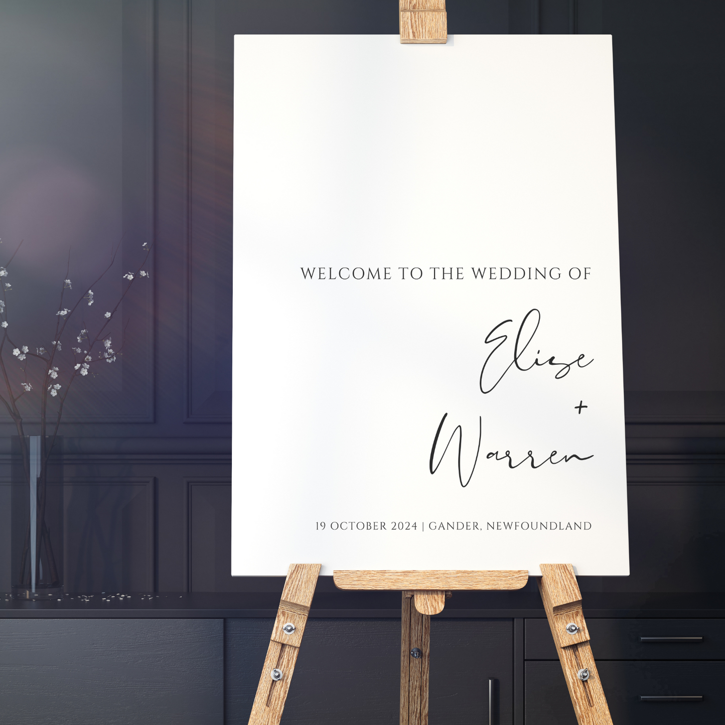 A minimalist wedding welcome sign sits on an easel in a dark-painted room.