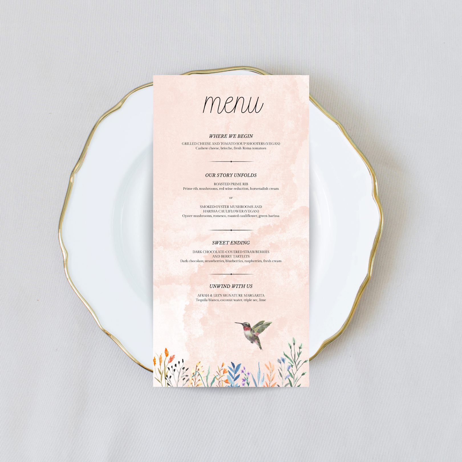 A light pink wedding menu with flowers and a hummingbird at the bottom sits on a white plate with gold trim against a cream coloured background.