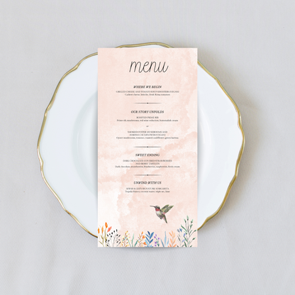 A light pink wedding menu with flowers and a hummingbird at the bottom sits on a white plate with gold trim against a cream coloured background.