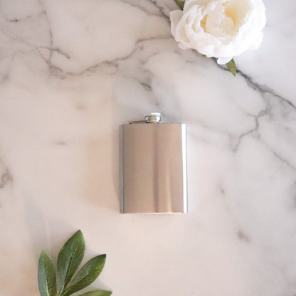 A plain stainless steel flask is pictured on a white marble background with a white flower towards the top and a leaf peaking out at the bottom of the image.