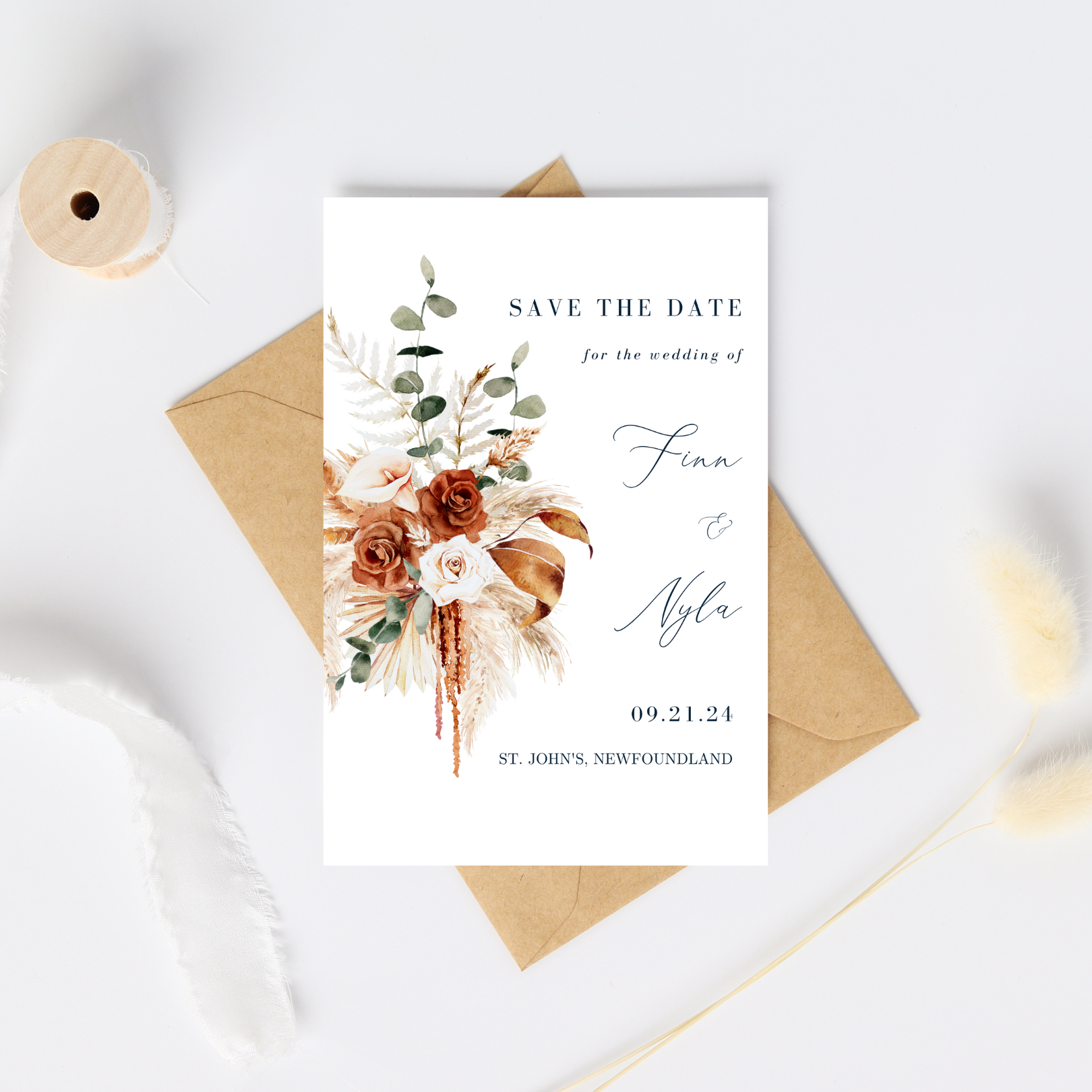 A white card with an image of a bohemian style floral bouquet sits on top of a kraft paper envelope. A spool of partially unwound white ribbon frames the card. On the Save The Date, the guest information is positioned on the right hand side.