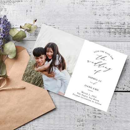 A card can be seen laying next to a kraft paper envelope and a sprig of lilac against a rustic wooden background. The card has a picture of a couple to the left and information for guests to the right.