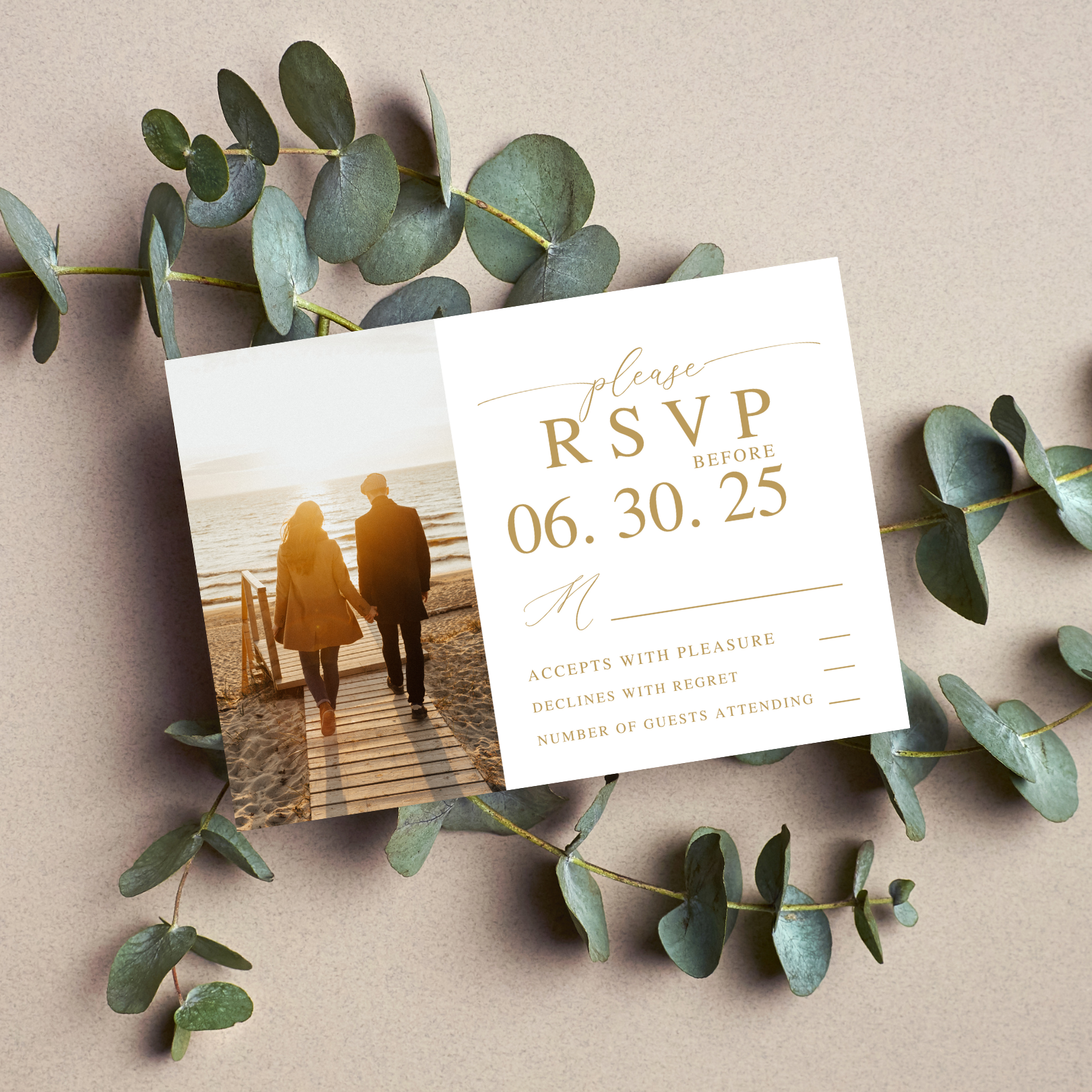 An R.S.V.P. card sits on top of a spray of eucalyptus on a neutral coloured background. The R.S.V.P. card has a photo of a couple on the left hand side who have their backs turned to the camera and are walking down the beach. On the right side of the card is information for guests.
