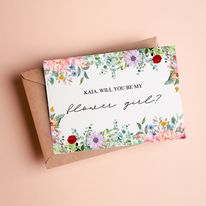 A card with a floral border lays on top of a kraft paper envelope against a light pink background with the phrase "Kaia, will you be my flower girl?".