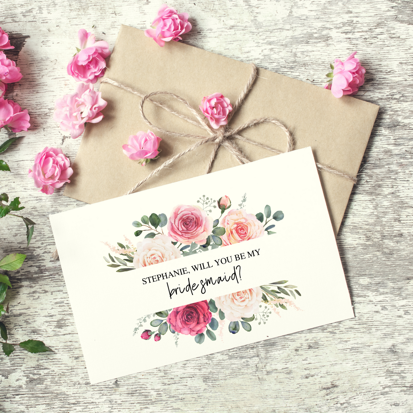 A white card with a burst of pink hued roses can be seen laying on top of a kraft paper envelope that's tied with a thin piece of brown string. There are small, pink roses sprinkled over the kraft envelope with a glimpse of a full bouquet peaking in from the left hand side of the image against a grey textured background.