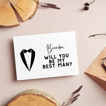 A white card with the silhouette of a tuxedo and the words "Brandon, will you be my best man?" can be seen against a light pink background with disks of wood and branches decorating the perimeter.