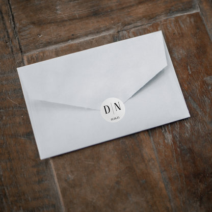 A white envelope lays on a dark wooden floor with a round sticker holding the flap closed. The white sticker has the initals "D" and "N" separated by a line with the date "07.26.25" below.