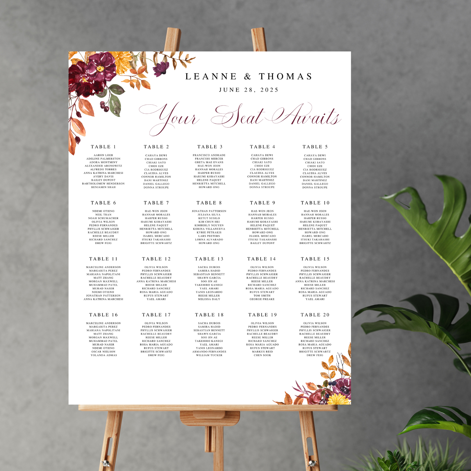 A seating chart sits on a wooden easel against a grey background with a monstera plant leaning in on the bottom right hand side of the image. The seating chart has a floral burst in the top left hand and bottom right hand corners and a list of tables in the middle. On the top, the chart reads "Leanne & Thomas...June 28, 2025... Your seat awaits".