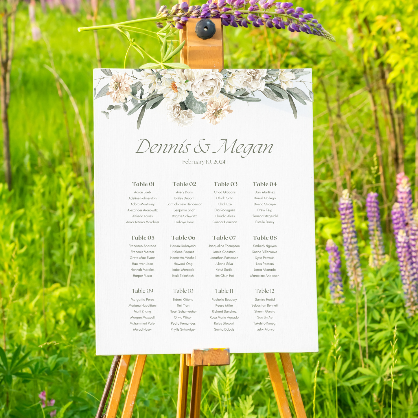 A seating chart sits on a wooden easel in a grassy field. The chart has a floral design on the top and a list of names below.