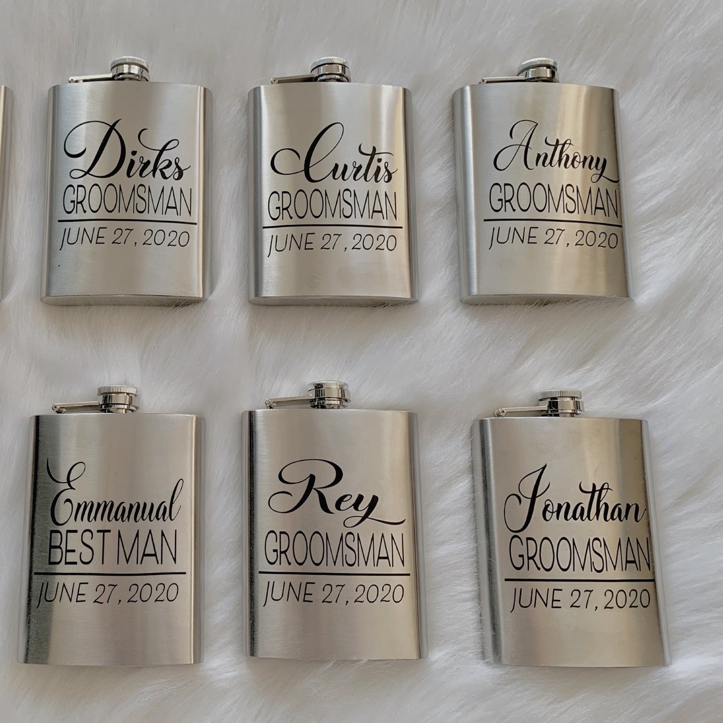 Six stainless steel flasks can be seen laying on a white background. Each flask has a name, title (i.e. groomsman) and a date.