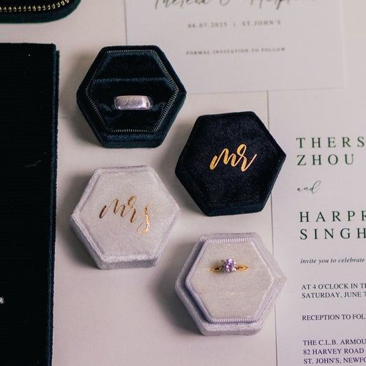 Black and white hexagon ring boxes lay open next to some invitations. The boxes have "mr." and "mrs." written on them in gold lettering. A ring sits in each box.
