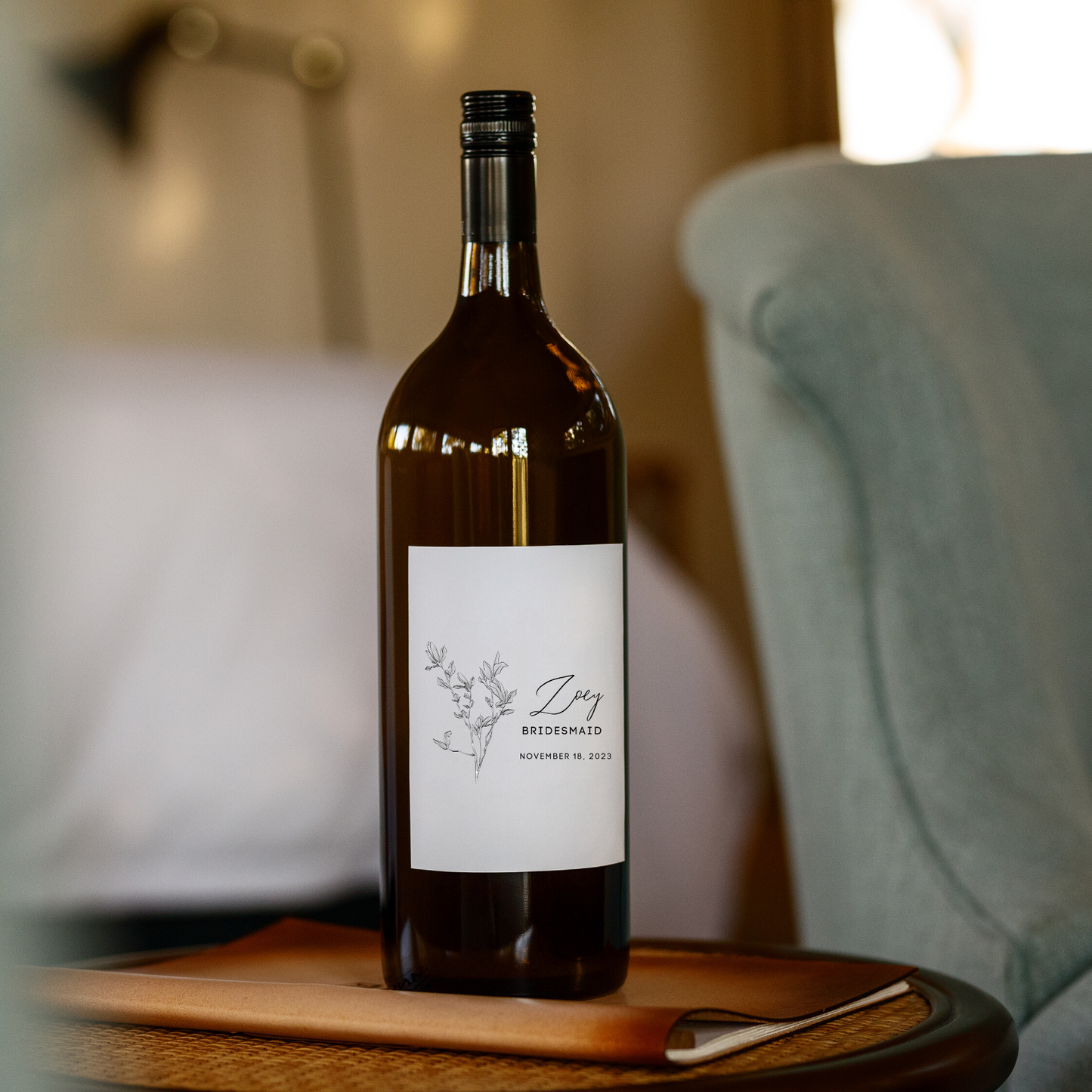A dark drown wine bottle sits on a wooden side table in a cozy living room. The label on the wine bottle has a minimalist flower design on the left and the words "Zoey", "Bridesmaid" and "November 18, 2023 to the right.