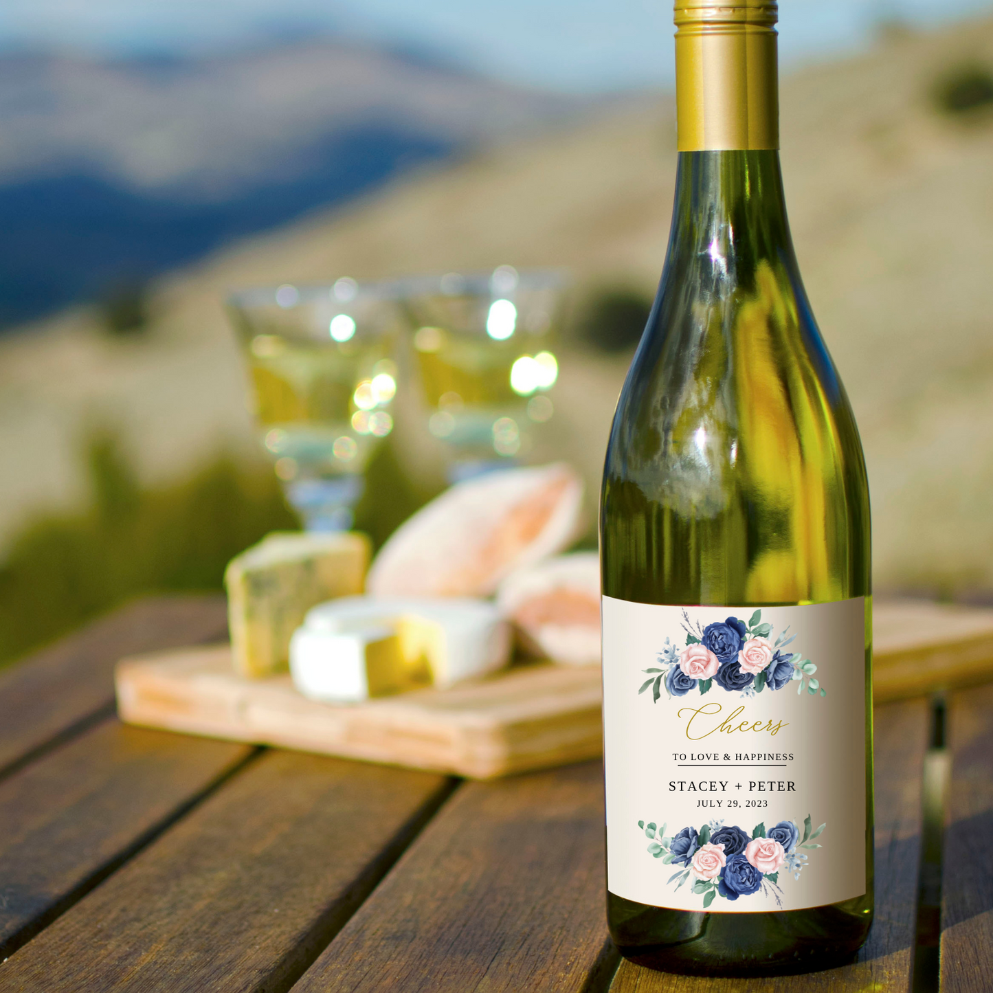 A bottle of wine sits on a wooden table at the edge of a cliff. Two glasses of wine and a cheese plate sit behind the bottle of wine. On the bottle's label, there are two clusters of blue and blush coloured roses: one on the top and one on the bottom. In between the flowers are the words "Cheers to love and happiness" and "Stacey + Peter... July 29, 2023".