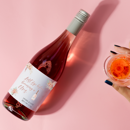 A bottle of rosé lays against a pin background. A hand appears from the lower right side of the image holding a glass of the rosé. A custom wine label with the words "Last sip before becoming a Mrs." and "Denise & Levi...July 6th, 2024" can be seen on the bottle.