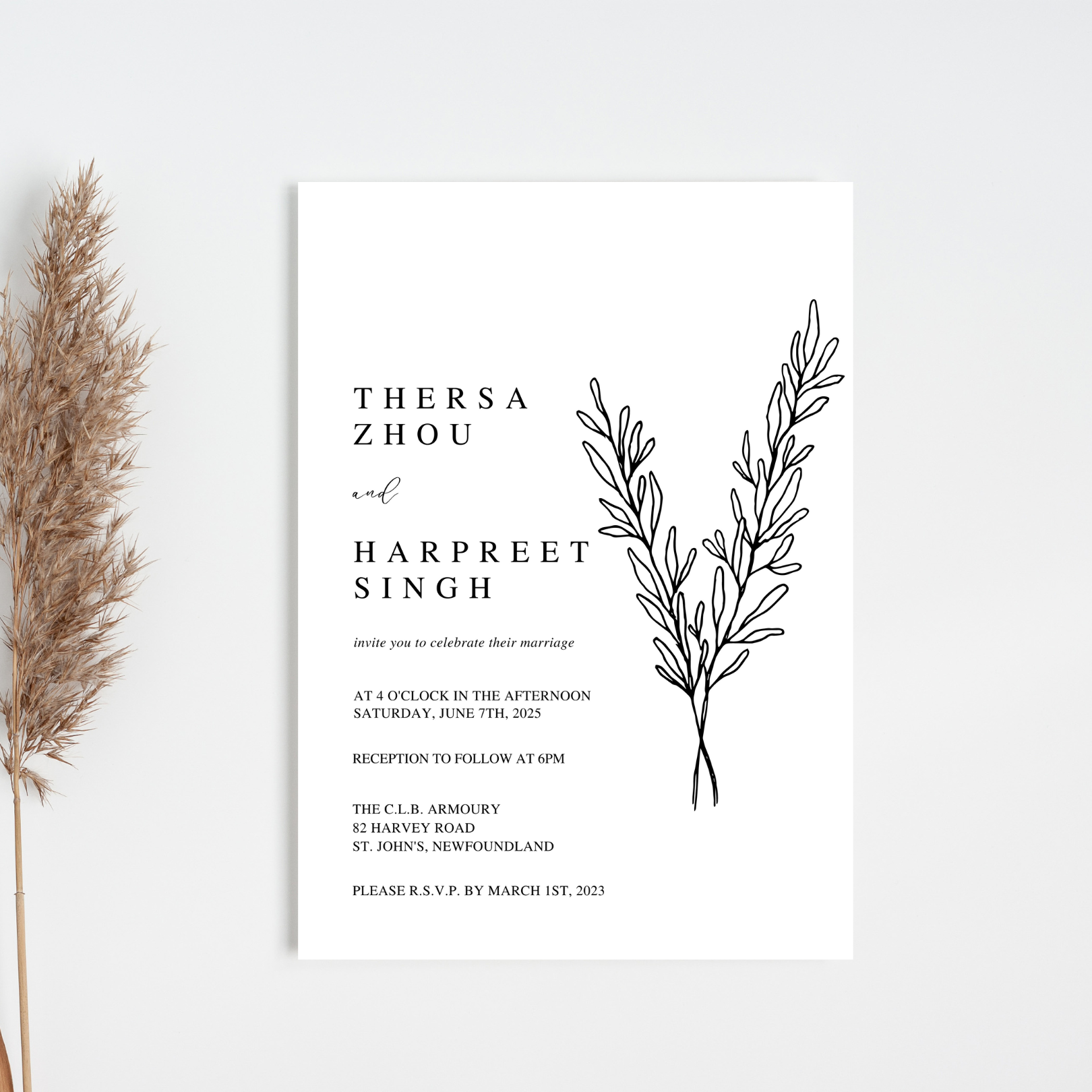 A white, minimalist wedding invitation sits against an off-white background. A piece of pampas grass can be seen peaking out of the left-hand side of the photo.