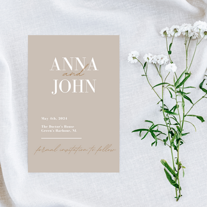 A beige coloured card lays on a white cotton cloth next to a small cluster of white flowers. The Save The Date card reads "Anna and John... May 4th, 2024... The Doctor's House... Green's Harbour, NL.... formal invitation to follow" 