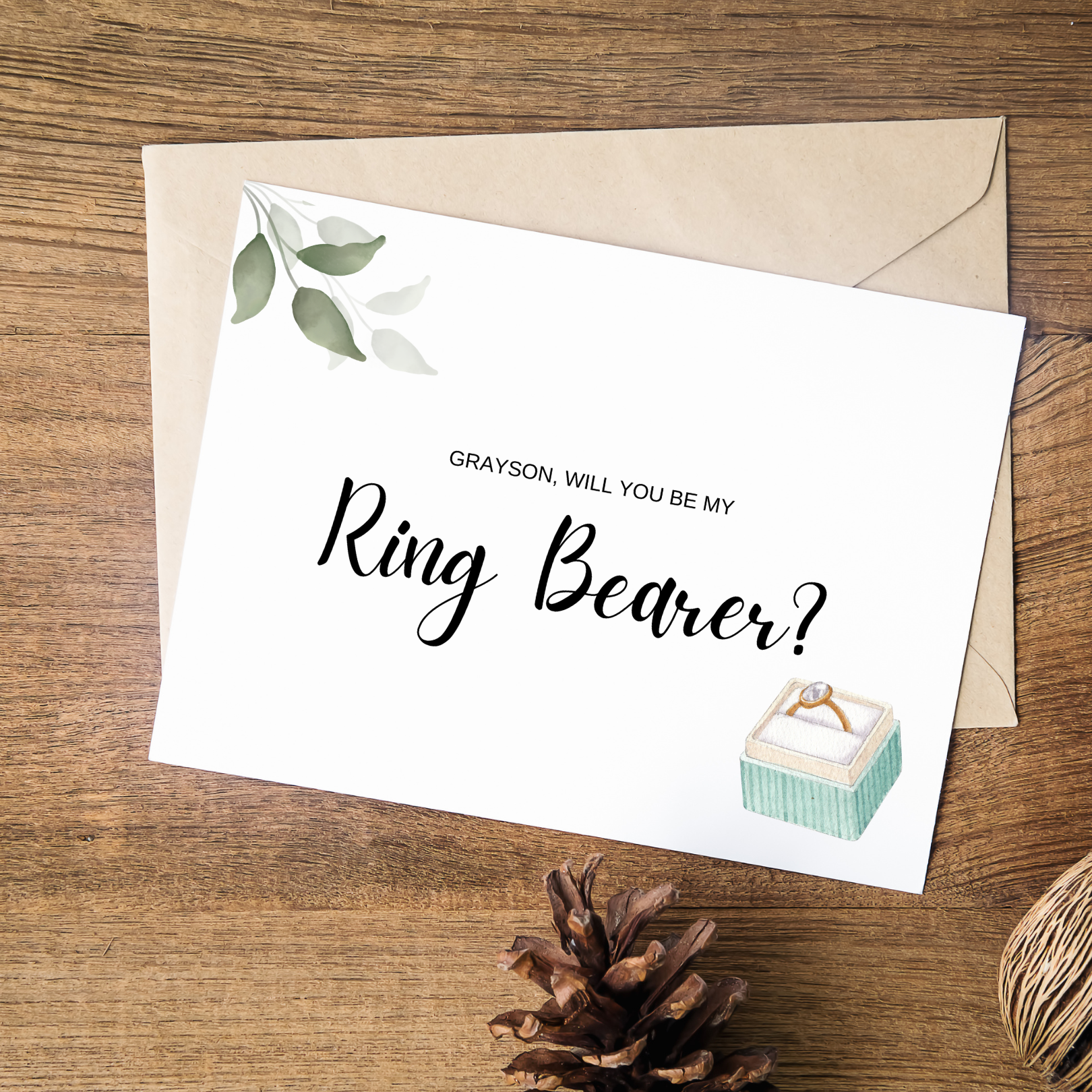 A white card can be seen with olive green leaves in the top left hand corner and a square ring box in the bottom right hand corner. The card says "Grayson, will you be my ring bearer?" and sits on top of a beige coloured envelope against a medium wood background. Below the card, there is a pine cone.